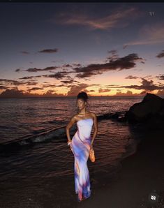 a woman standing on top of a beach next to the ocean at sunset with her hands in her pockets