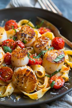 scallops and tomatoes are on top of pasta in a black plate with silverware