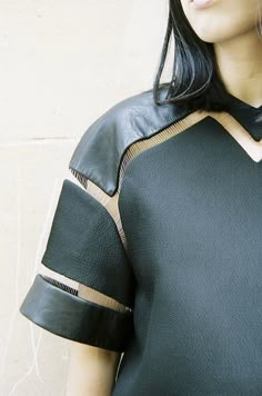 a woman with black hair wearing a green top and brown leather sleeves, standing against a wall