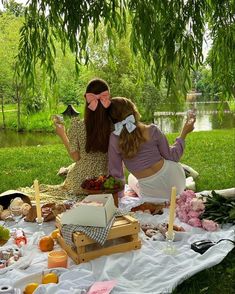 two women sitting on a blanket in front of a tree eating food and drinking wine