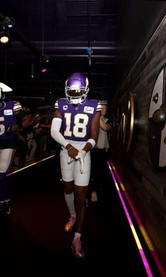 two football players are walking down the tunnel with their helmets on and one is wearing a purple uniform