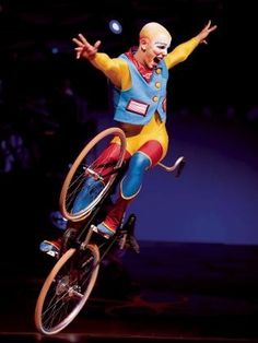 a clown riding a bicycle in the air with his arms out and legs spread wide