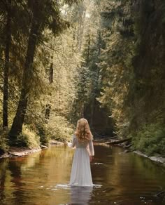 a woman in a white dress is standing in the middle of a river surrounded by trees