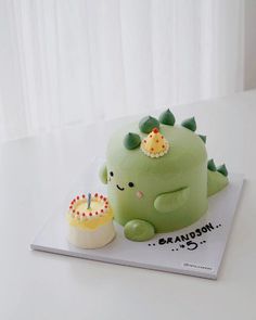 a green dinosaur cake sitting on top of a white table next to a cupcake