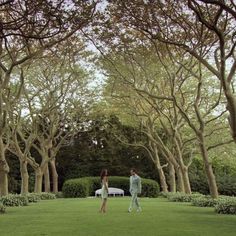 two people walking in the middle of a lush green park with trees and benches behind them