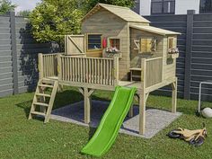 a wooden play house with a green slide in the grass next to it and a gray fence