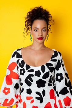 a woman with curly hair standing in front of a yellow wall wearing an animal print top