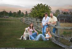 three people and a dog are sitting on the grass near a fence in an open field