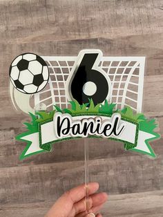 a hand holding up a cake topper with a soccer ball and goal on it