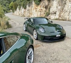 two green sports cars parked next to each other on the side of a mountain road