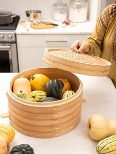 a woman holding a wooden basket filled with squash and gourds on top of a kitchen counter