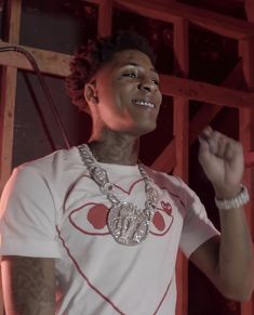 a young man wearing a white t - shirt and silver necklace standing in front of a wooden structure