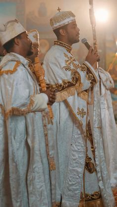 two men dressed in white and gold standing next to each other while holding a microphone