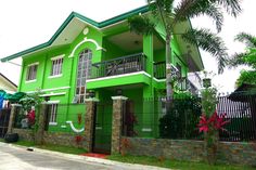 a green house with black iron fence and trees in the front yard, on a sunny day