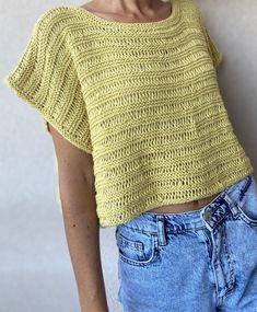 a woman with short hair wearing a yellow knitted top and jeans, standing against a white wall