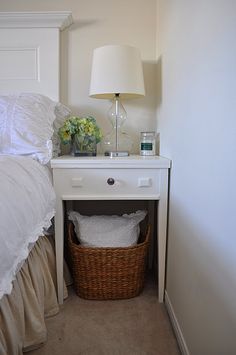 a white bed with a basket under it and a lamp on the nightstand next to it