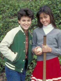 two young people standing next to each other in front of some bushes and trees, one holding a baseball bat