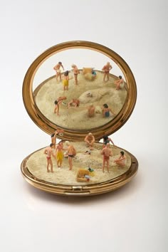 miniature people playing in the sand inside an open gold compact case on a white surface