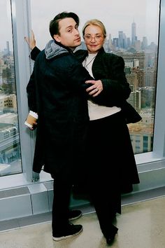 a man and woman standing next to each other in front of a window looking out at the city