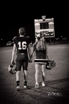 a man and woman in football uniforms holding hands on a field at night with the scoreboard in the background