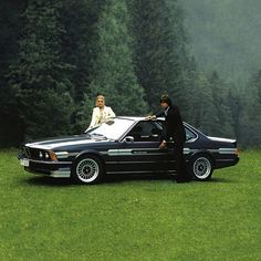 two people standing next to a black car in the middle of a field with trees