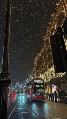 a red double decker bus driving down a city street at night with snow falling on the ground