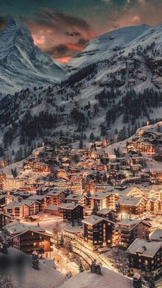 a snowy mountain town is lit up at night with lights on the buildings and mountains in the background