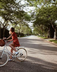 a woman in a red dress riding a white bike down a street with trees on both sides