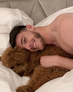 a shirtless man laying in bed with his brown dog and smiling at the camera