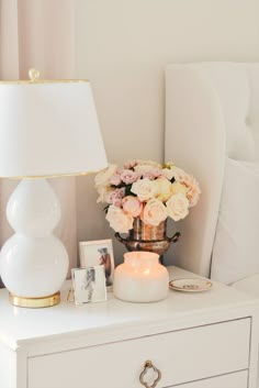 a white dresser topped with a vase filled with flowers next to a lamp and pictures