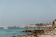 people are swimming in the water near some rocks and boats on the shore, with buildings in the background