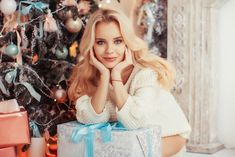 a beautiful blonde woman sitting next to a christmas tree holding a present box with her hand