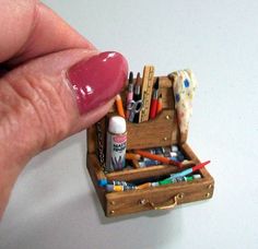 a miniature wooden box with pens, pencils and markers in it is being held by someone's finger