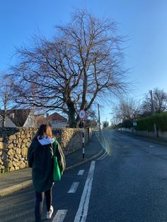 a woman walking down the street in front of a stone wall and tree with no leaves