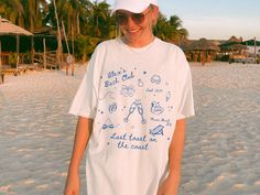 a woman standing on top of a sandy beach next to the ocean wearing a white hat and t - shirt