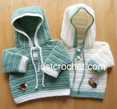 two crocheted sweaters with hoodies are shown on a wooden surface, one is green and the other is white