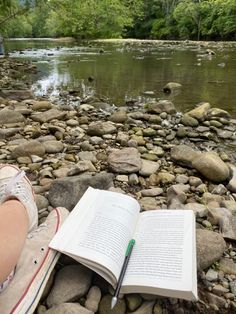 a person is sitting on rocks with an open book in front of them and the river behind them