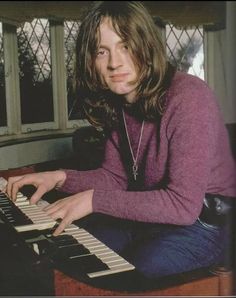 a man with long hair sitting at a keyboard in front of a window and looking into the camera