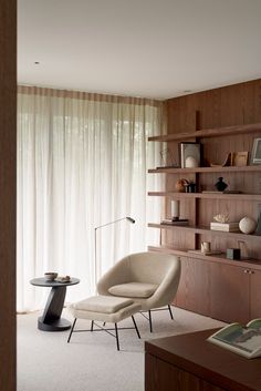 a living room filled with furniture next to a wall mounted book shelf and window covered in sheer curtains