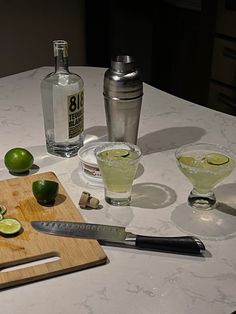 limes are sitting on the table next to two glasses and a cutting board with alcohol