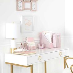 a white desk topped with lots of pink items next to a lamp and pictures on the wall