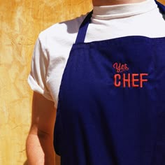 a man wearing an apron with the word chef embroidered on it's chest, standing in front of a wooden wall