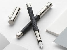 three fountain pens sitting on top of a white table next to an open notebook and pen