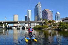 a man is standing on a paddle boat in the water with tall buildings behind him