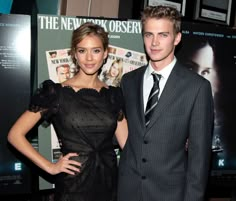 a man and woman standing next to each other in front of a wall with posters on it