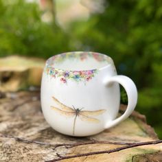 a coffee cup with dragonflys painted on it sitting on a tree stump in the woods