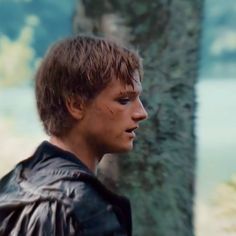 a young man wearing a leather jacket standing in front of a tree and looking off into the distance