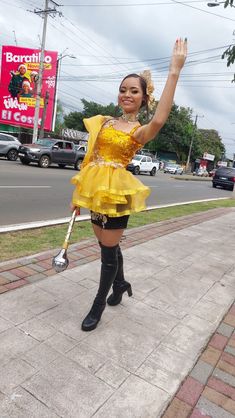 a woman in a yellow dress and black boots is standing on the sidewalk with her arms up