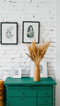 a green dresser with two pictures on the wall and some dried grass in a vase