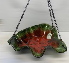 a red and green leaf shaped bowl hanging from chains on a white wall with a tag attached to it